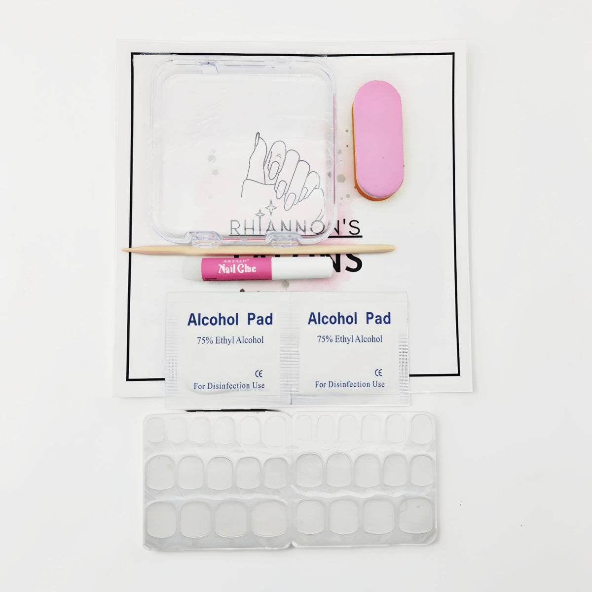 Nail kit contents laid out on a white printed card over a speckled gray countertop, including a clear plastic tray, a pink nail file, a wooden stick, a pink tube labeled Nail Glide, two Alcohol Pad packets, and a sheet of adhesive nail tabs.