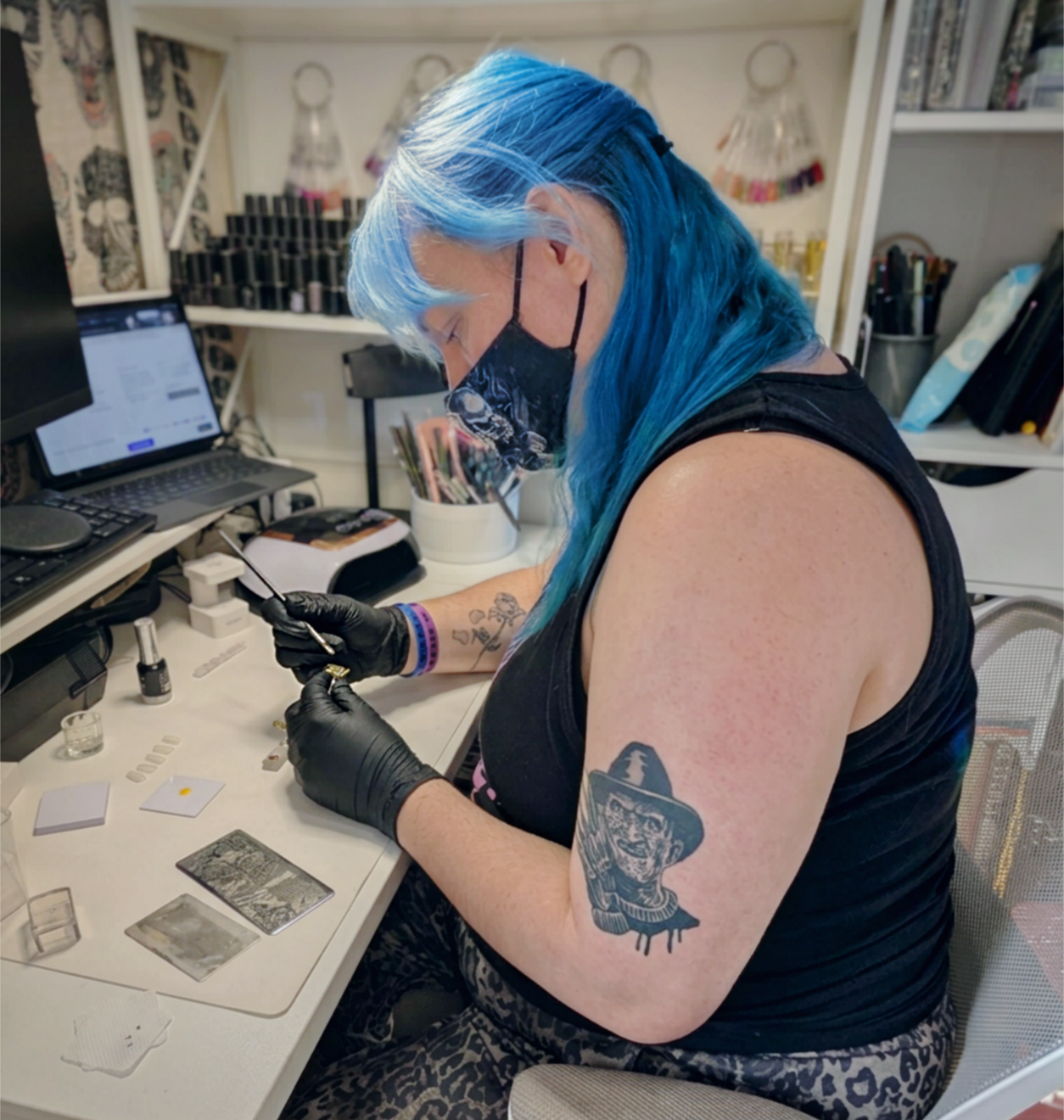 A nail artist with bright blue hair sits at a white desk in a nail studio, wearing black gloves and a black sleeveless top while working on a set of nails. 